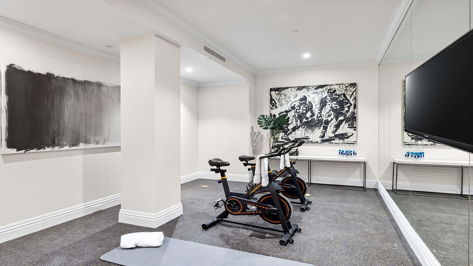 This is a well-appointed home gym featuring two stationary bikes, a gray exercise mat, and modern artwork. The room has a neutral color palette with white walls and gray flooring, creating a clean and inviting space. A large mirror on one wall enhances the sense of space and allows for form checks during workouts.