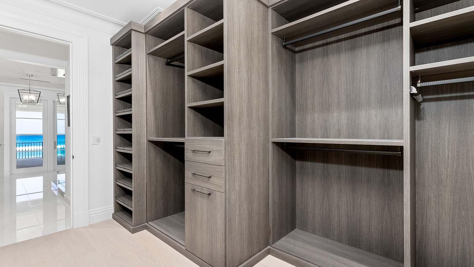 This is a well-organized walk-in closet featuring custom wood shelving and drawers. The closet is spacious and offers ample storage for clothing and accessories. A glimpse of the ocean view through a nearby doorway adds a luxurious touch.