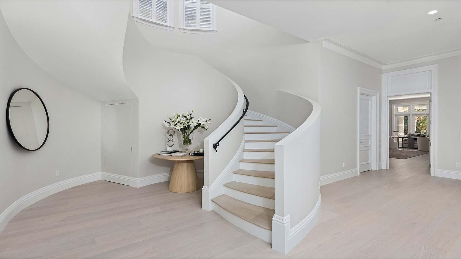 This is an interior shot of a bright and elegant hallway featuring a curved staircase. The walls are painted in a neutral tone, complemented by light hardwood flooring. A round mirror and a small round table with floral arrangements add to the sophisticated ambiance, while the staircase with its white banister and black handrail serves as a focal point.