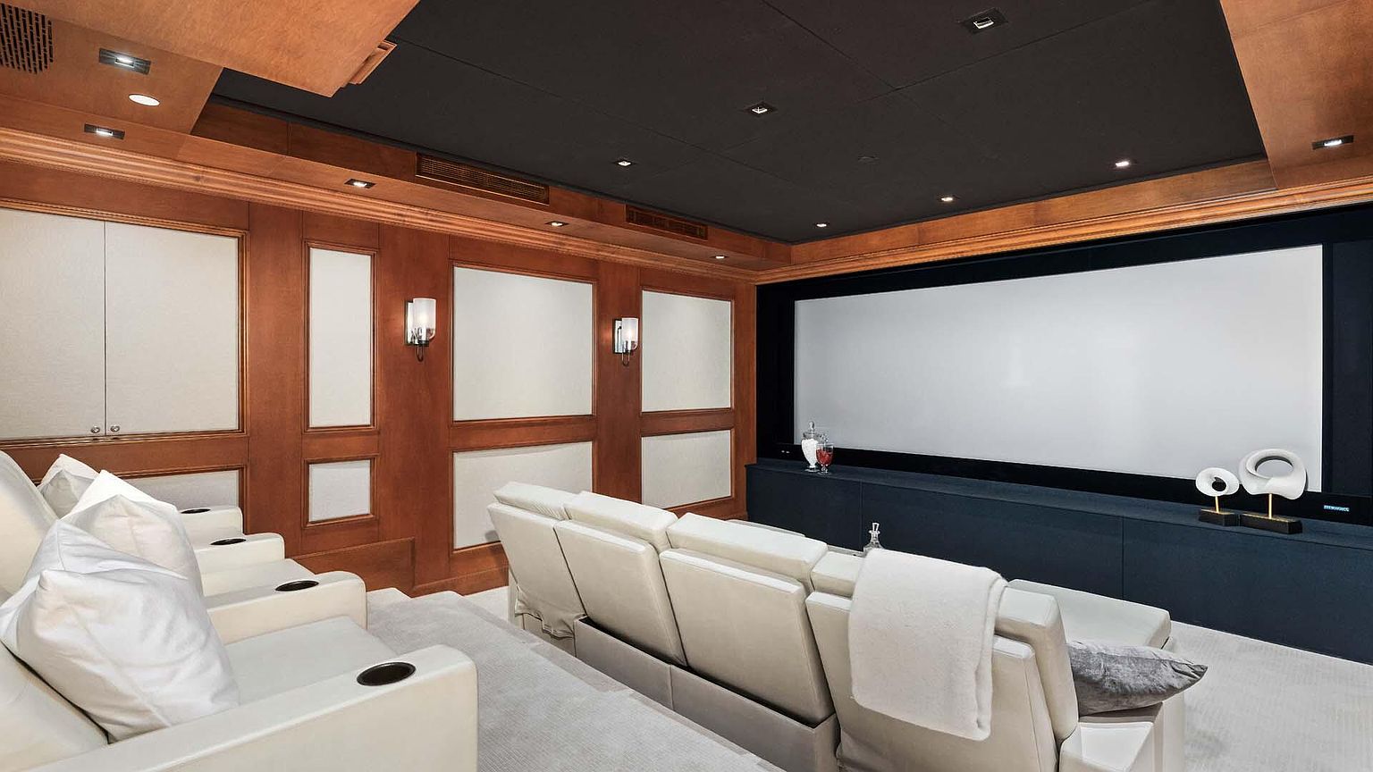 This is an interior shot of a home theater. The room features tiered seating with plush, white leather recliners, a large projection screen, and wood-paneled walls with sconce lighting. The ceiling is dark, creating an immersive viewing experience. The overall impression is one of luxury and comfort.