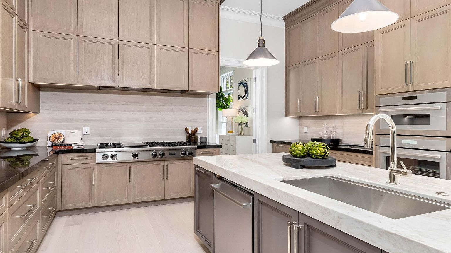 This is a well-lit kitchen featuring light wood cabinetry, stainless steel appliances, and a marble countertop island with a sink. The kitchen has a modern design with a neutral color palette, creating a clean and sophisticated atmosphere. The perspective is from a medium shot, showcasing the island and the cooking area.
