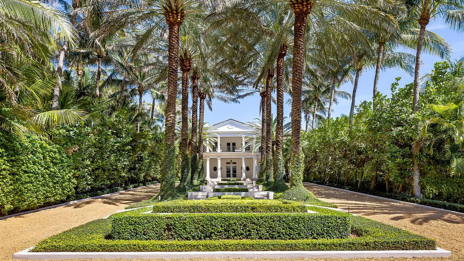 This is a stunning front view of a grand estate, showcasing a symmetrical two-story white house framed by towering palm trees and meticulously manicured greenery. The driveway leads directly to the house, emphasizing the property's impressive scale and luxurious landscaping. The overall impression is one of elegance and privacy.
