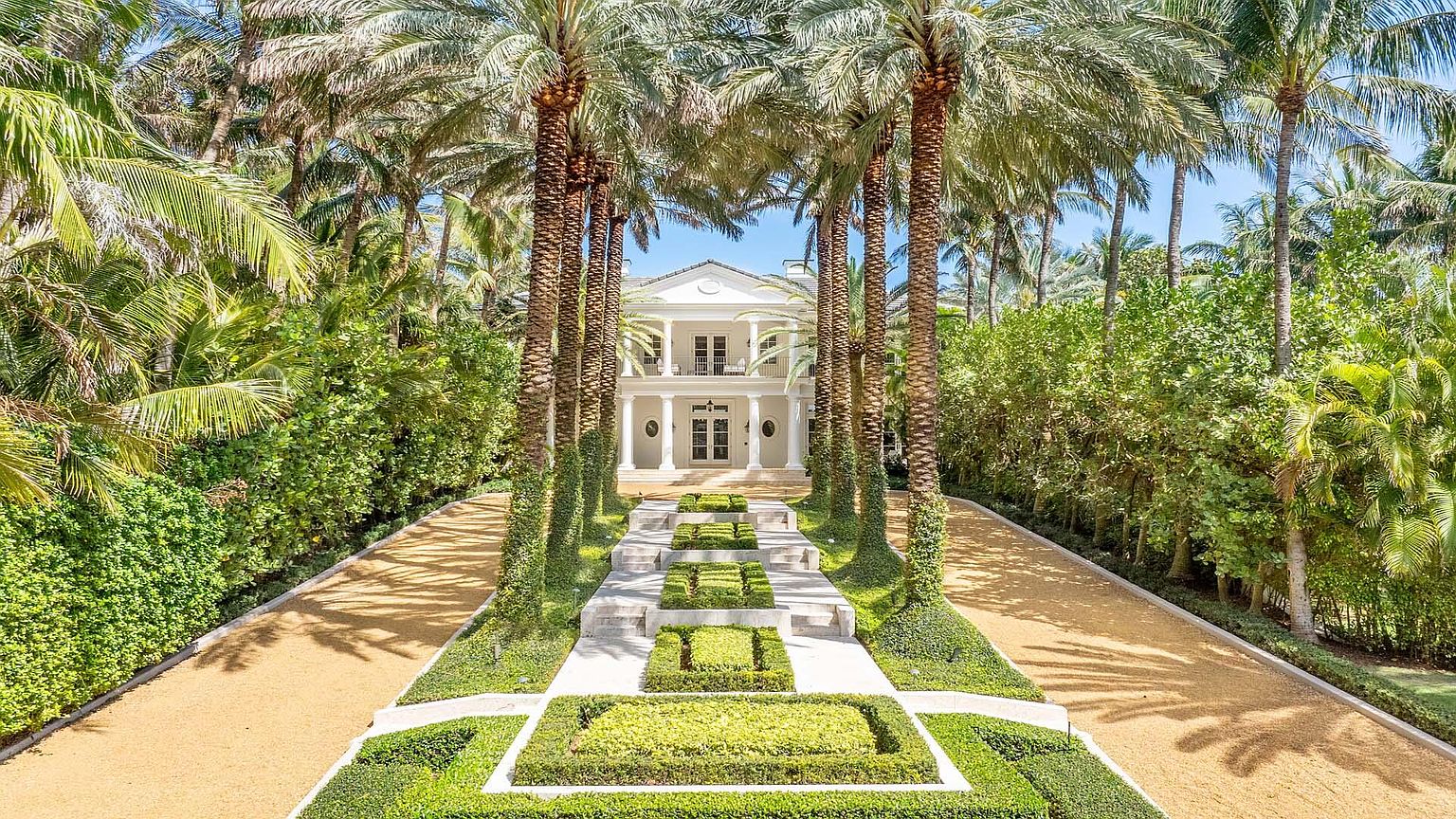 This is a stunning front view of a grand estate, showcasing a symmetrical facade with white columns and a second-story balcony. The property is framed by rows of tall palm trees and meticulously manicured hedges, leading to a long, gravel driveway. The overall impression is one of luxury, privacy, and classic elegance.
