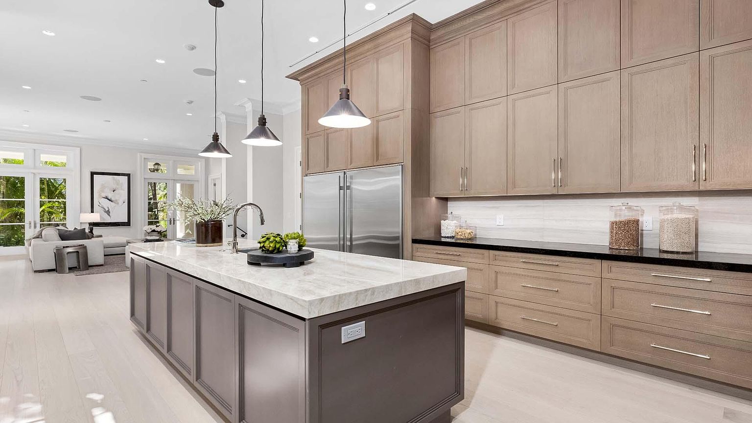 This is a bright and spacious kitchen featuring a large center island with a marble countertop and dark gray cabinetry. The kitchen also includes stainless steel appliances, light wood cabinets, and a black countertop along the back wall. The open floor plan leads into a living area, creating a seamless flow, and the perspective is from a wide angle to capture the entire space.