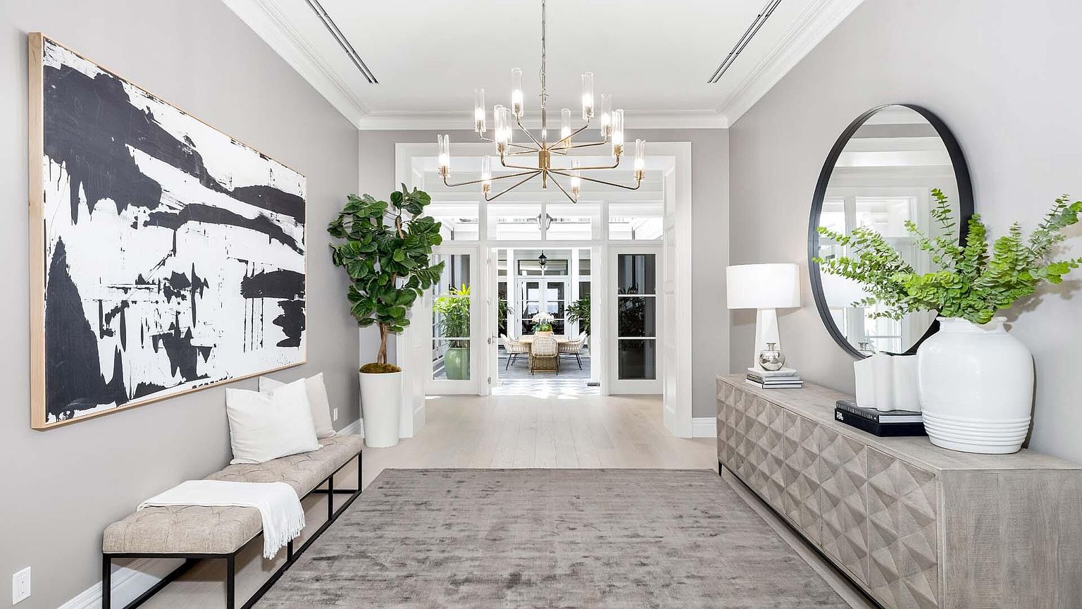 This is an interior shot of a grand hallway featuring a neutral color palette with gray walls and light wood floors. The space is decorated with a large abstract painting, a bench with pillows, a console table with a round mirror and decorative vases, and a large potted plant. A modern chandelier hangs from the ceiling, and a doorway leads to another room with a dining table visible in the background, creating a sense of depth and openness.