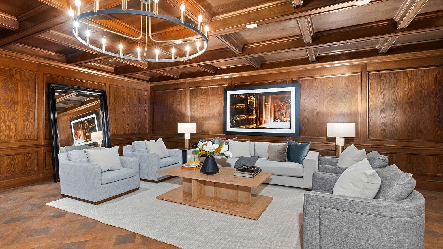 This is an interior shot of a luxurious living room featuring rich wood paneling and a coffered ceiling with a modern chandelier. The room is furnished with comfortable gray armchairs and a sofa, arranged around a light-colored area rug and a wooden coffee table. A large framed artwork hangs on the wall, complemented by table lamps, creating a warm and inviting atmosphere.