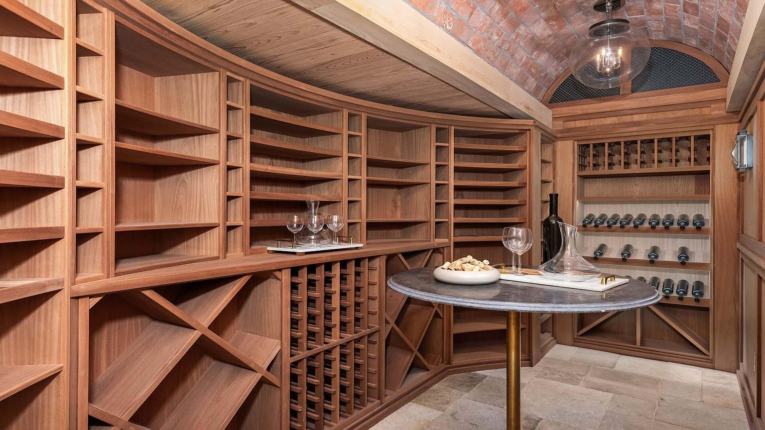 This is a well-appointed wine cellar featuring custom wooden shelving designed to hold numerous bottles. A round table with a marble top and gold base sits in the center, displaying wine glasses and a decanter, suggesting a tasting area. The brick arched ceiling and stone flooring add to the cellar's sophisticated and classic ambiance.