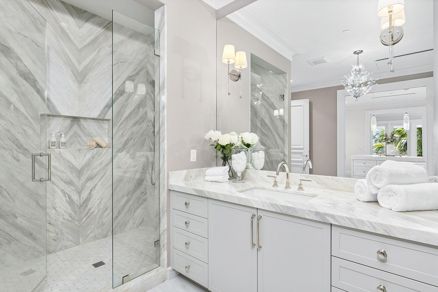 This is a luxurious primary bathroom featuring a glass-enclosed shower with marble chevron-patterned walls and a built-in niche. The vanity has a marble countertop, light gray cabinetry with elegant hardware, and a large mirror reflecting a view into another area of the bathroom. The overall impression is clean, bright, and sophisticated.