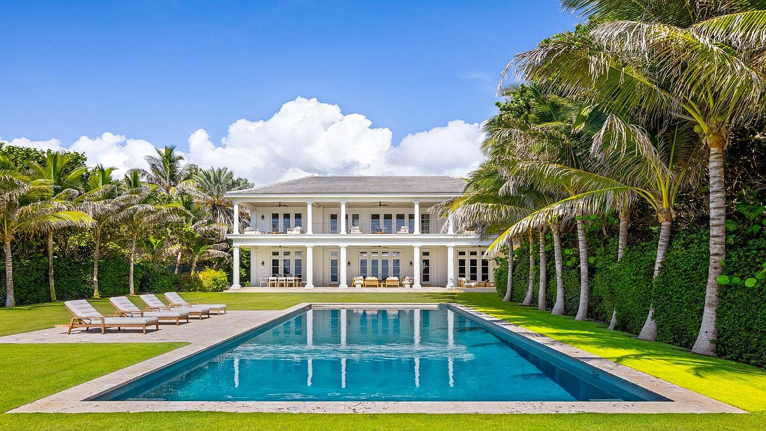 This image showcases a luxurious backyard featuring a rectangular swimming pool with crystal clear water reflecting the architecture of the two-story white house. The house has a colonial style with columns and balconies. The pool is surrounded by a well-manicured lawn and framed by palm trees, creating a serene and private oasis.