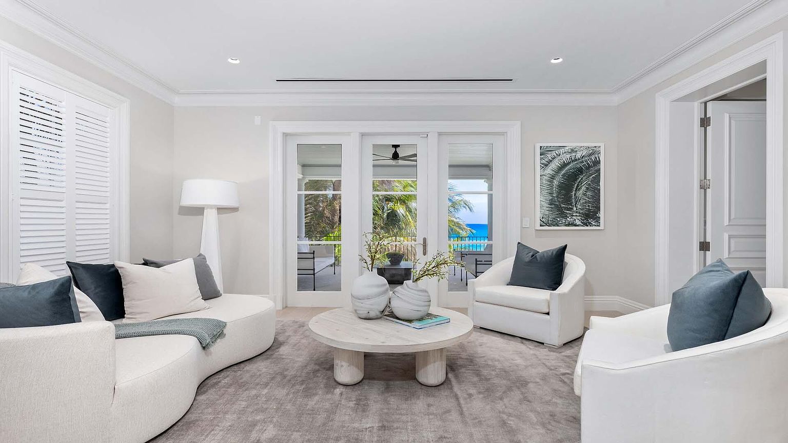 This is a bright and airy living room featuring a curved white sofa, two white armchairs, and a round coffee table. The room has large windows and doors that lead to an outdoor area with a view of the ocean. The overall style is modern and minimalist, creating a serene and inviting atmosphere.