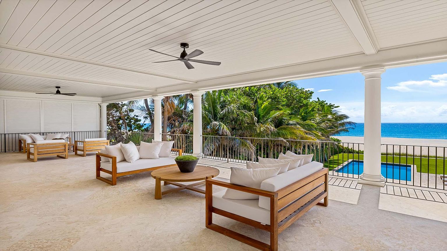 This image showcases a luxurious outdoor patio or balcony area with elegant wooden furniture and plush white cushions. The space overlooks a stunning ocean view, complete with a pool and lush greenery. The architectural details, including the white columns and ceiling fans, add to the upscale ambiance, making it an ideal spot for relaxation and entertainment.
