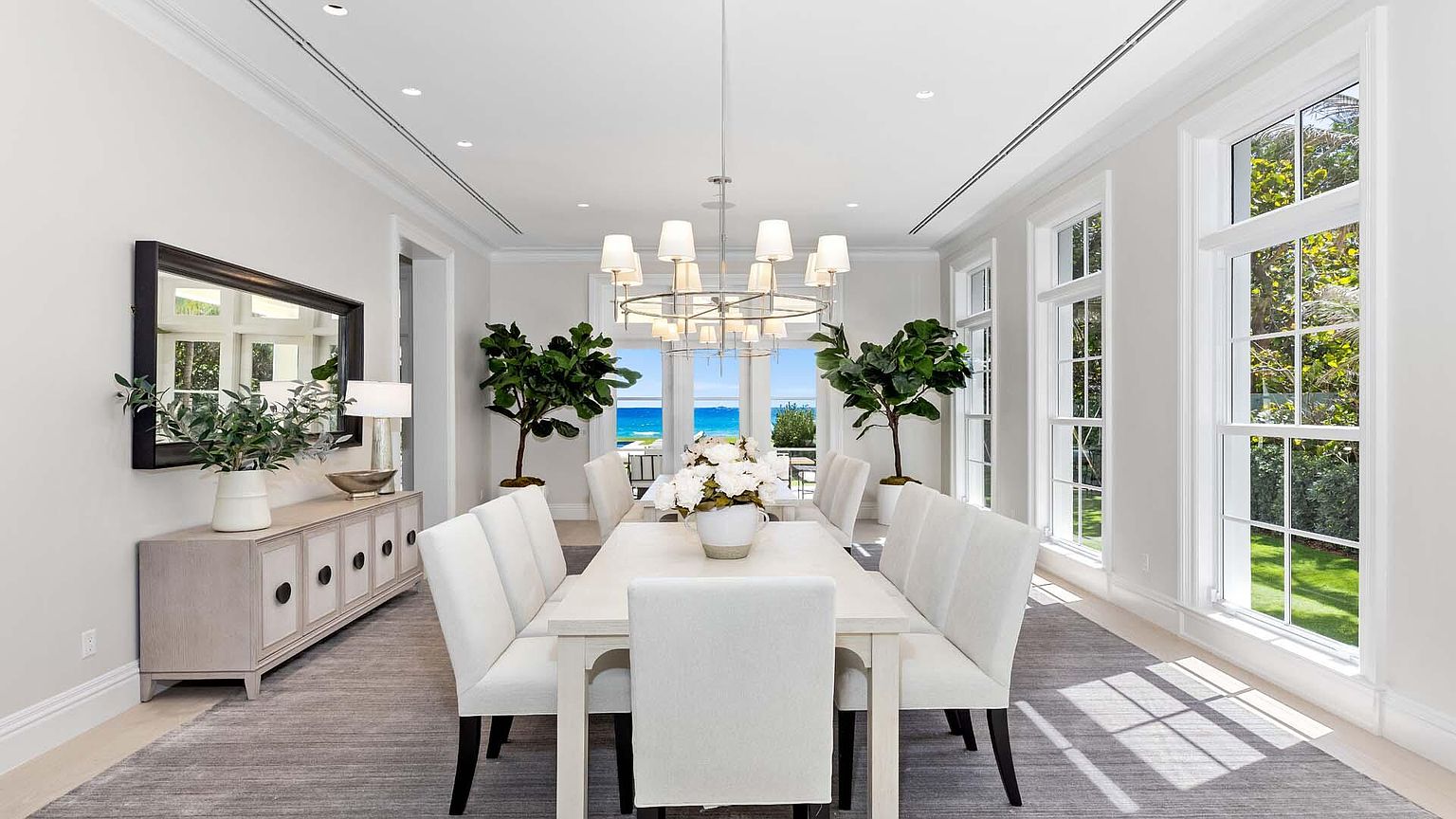 This is a bright and elegant dining room featuring a long white dining table with upholstered chairs, illuminated by a modern chandelier. Large windows offer a view of the ocean, enhancing the room's airy and luxurious feel. A sideboard with a mirror and decorative plants adds a touch of sophistication.