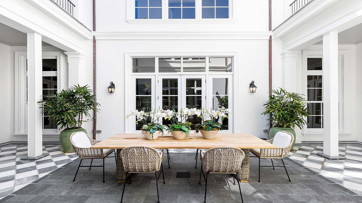 This image showcases an elegant outdoor patio area, featuring a wooden dining table surrounded by woven chairs with black metal legs. The patio is enclosed by a white building with large windows and columns, creating a courtyard effect. Potted plants add a touch of greenery, and the floor is paved with gray and white tiles in a checkered pattern, enhancing the sophisticated ambiance.