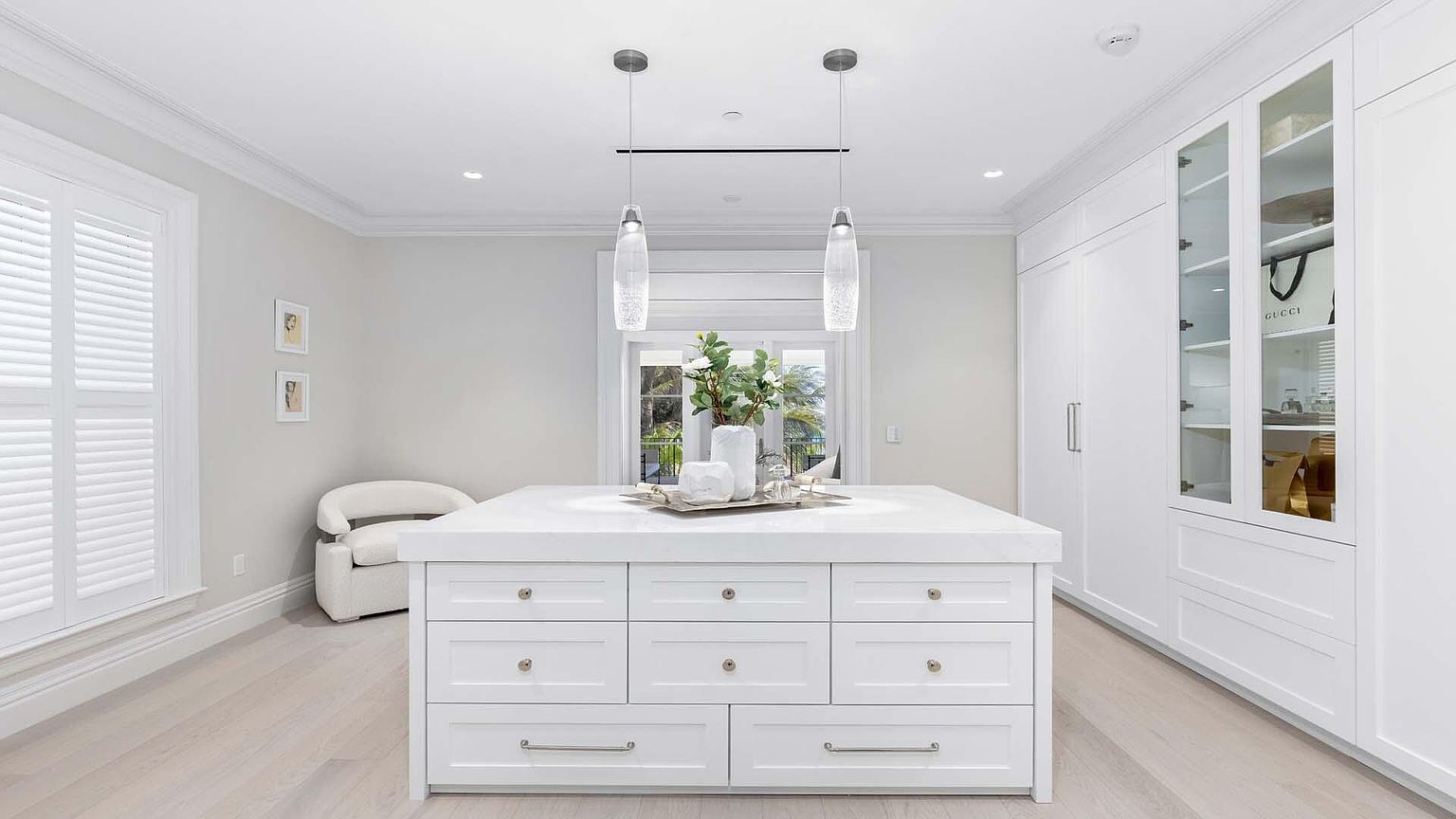 This is a bright and luxurious walk-in closet featuring custom white cabinetry with drawers and glass-fronted display cabinets. A central island with a white countertop provides additional storage and workspace. The room is well-lit with pendant lights and recessed lighting, creating an elegant and organized space.