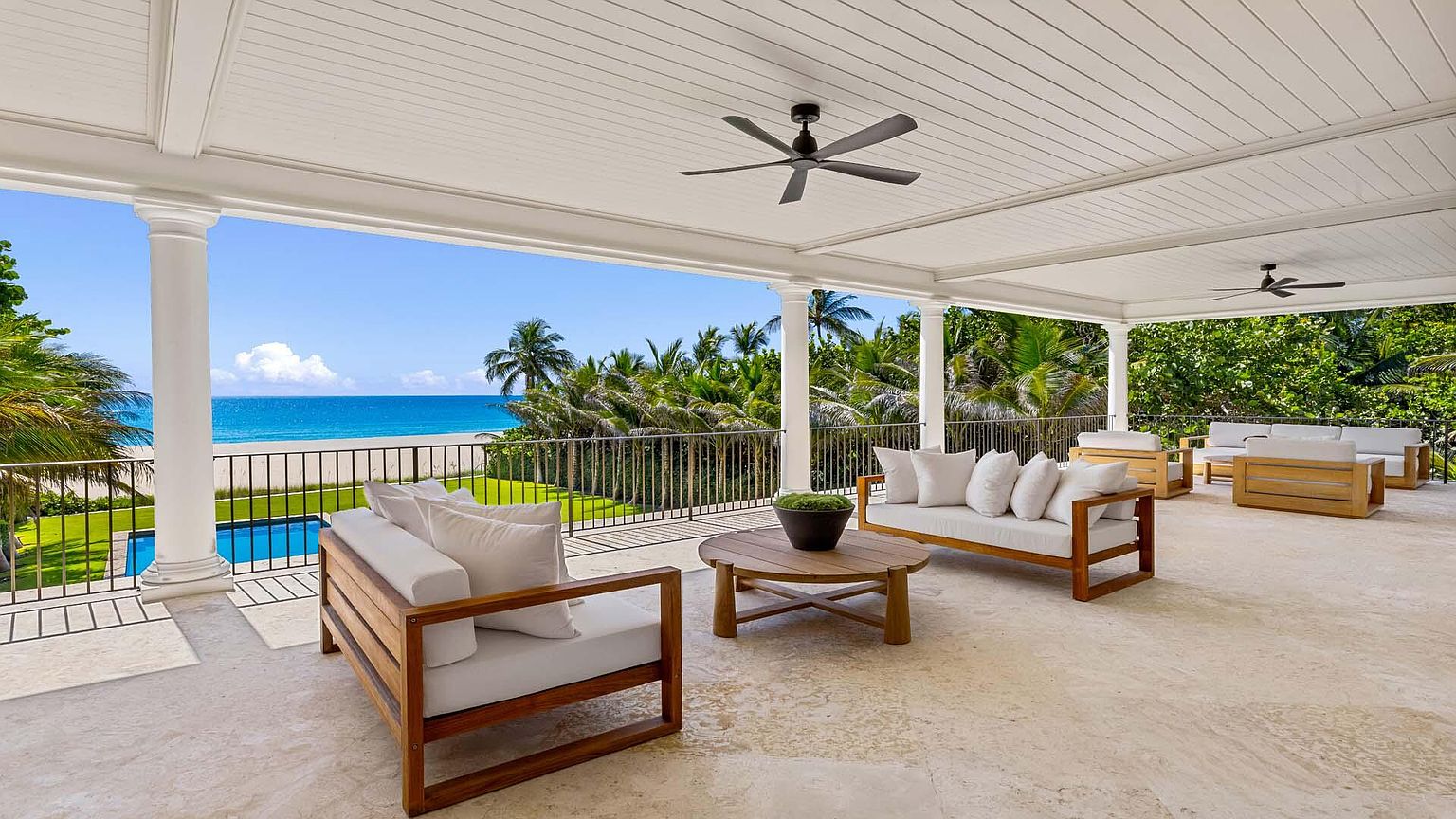This image showcases a luxurious outdoor patio or balcony area with elegant wooden furniture and plush white cushions. The space is covered by a white ceiling with ceiling fans and supported by classic white columns, offering stunning views of the ocean, beach, and lush greenery. The overall impression is one of sophisticated relaxation and upscale coastal living.