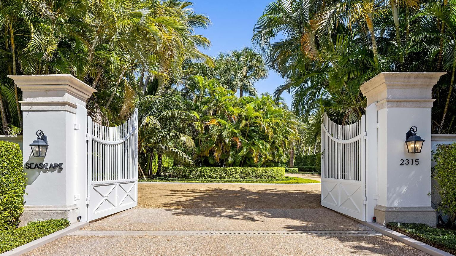 This image showcases a grand entryway to a property, featuring an open white gate flanked by substantial white pillars. Lush tropical landscaping, including palm trees and manicured hedges, creates a sense of privacy and luxury. The driveway is gravel, adding to the estate's upscale appeal.