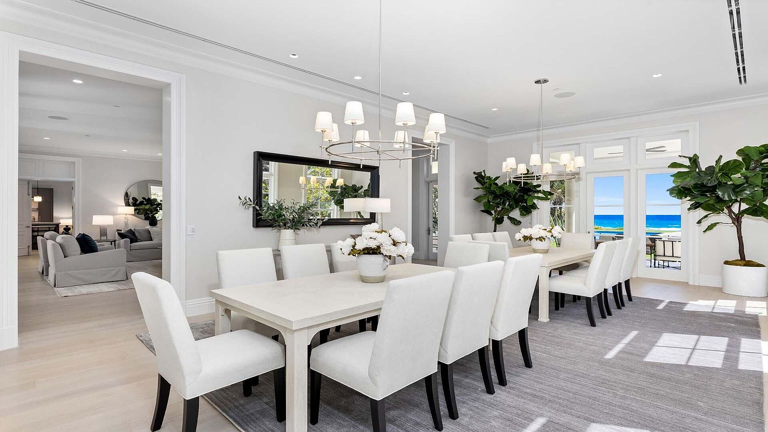 This is a bright and airy dining room featuring a long white dining table with seating for ten, illuminated by modern chandeliers. The room is decorated in neutral tones with a large mirror on the wall and a view of the ocean through the doors leading to a balcony. The overall impression is elegant and sophisticated, perfect for entertaining.