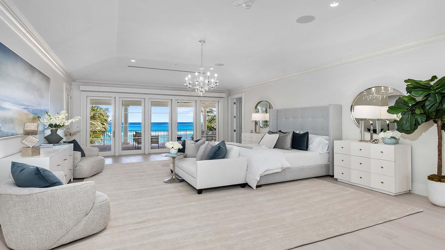 This is a luxurious primary bedroom featuring a king-size bed with a light gray upholstered headboard and a white duvet. The room has a serene atmosphere with a neutral color palette, complemented by a crystal chandelier and large windows offering ocean views. The space includes two armchairs, side tables, and dressers, enhancing the room's functionality and elegance.