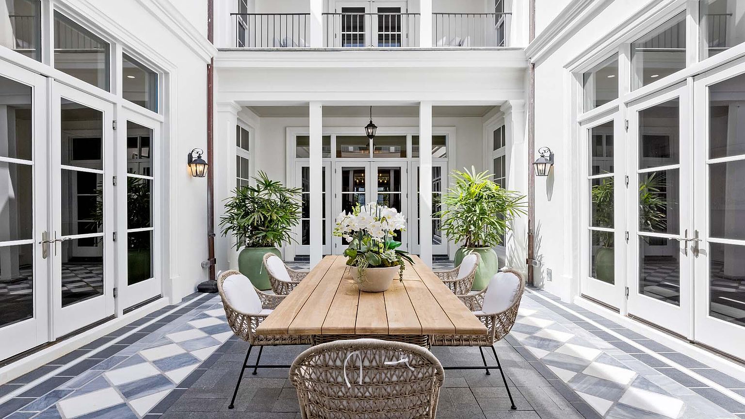 This image showcases a beautifully designed outdoor patio area, featuring a large wooden dining table surrounded by wicker chairs with white cushions. The patio is adorned with potted plants, adding a touch of greenery, and the flooring consists of a stylish checkered pattern. The space is framed by white architectural elements, including columns and French doors, creating an elegant and inviting atmosphere.