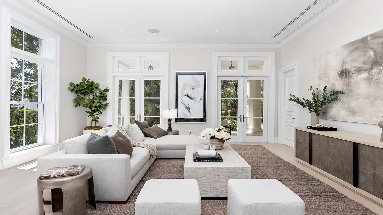 This is a bright and airy living room featuring a large white sectional sofa, a textured coffee table, and two matching ottomans. The room is decorated in neutral tones with pops of greenery from plants and artwork. Large windows and glass doors provide ample natural light and views of the outdoors, enhancing the sense of spaciousness and luxury.
