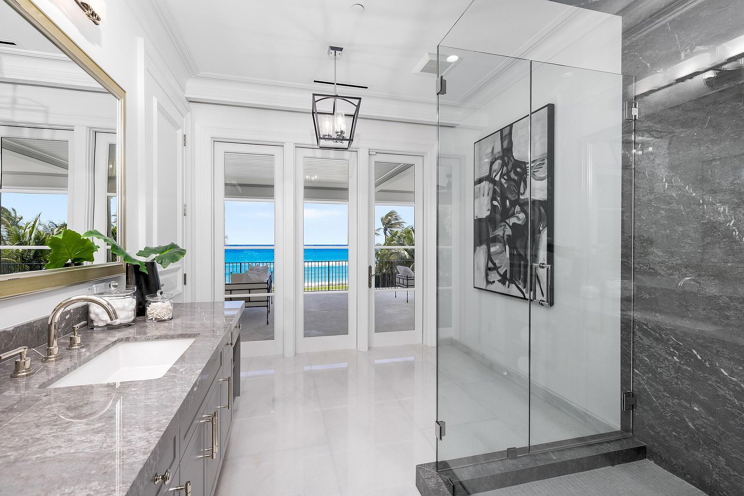 This is a luxurious primary bathroom featuring a gray marble countertop with dual sinks, a large mirror, and modern fixtures. The bathroom has a glass-enclosed shower with gray marble walls and a piece of art on the wall. The room opens to a balcony with ocean views, enhancing the spa-like atmosphere.