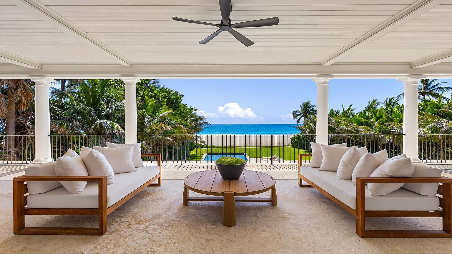 This image showcases a luxurious outdoor patio or balcony area with stunning ocean views. The space is furnished with two wooden-framed sofas adorned with white cushions, flanking a round wooden coffee table with a potted plant. The patio is covered with a white ceiling and supported by white columns, creating an elegant and inviting outdoor living space.
