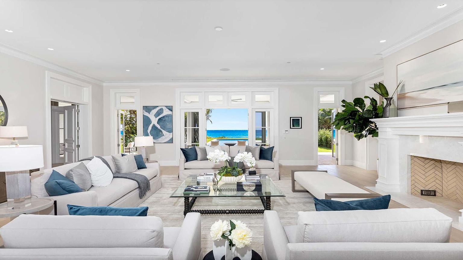 This is a bright and airy living room featuring two sofas, two armchairs, and a glass coffee table. The room is decorated in neutral tones with blue accents, and large windows offer a view of the ocean. A fireplace with a white mantel and a large piece of artwork add to the elegance of the space. The perspective is from the back of the room, looking towards the ocean view.