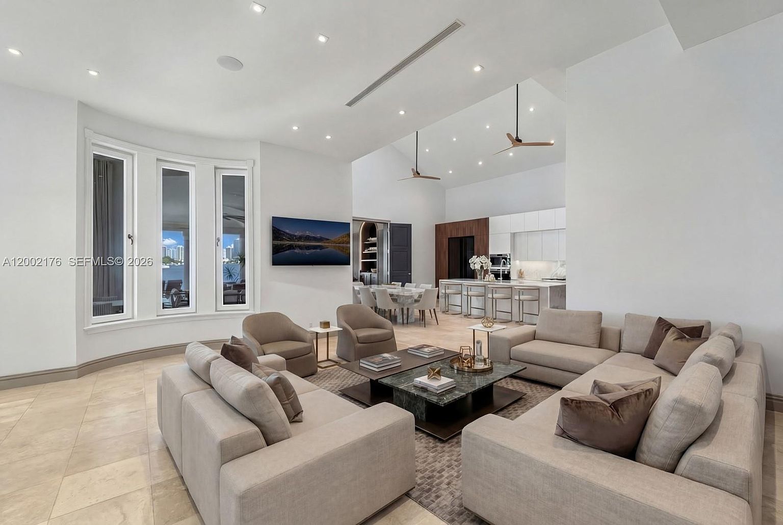 This spacious, open-concept living area features a neutral color palette with plush beige sofas arranged around a central stone-topped coffee table. The room flows seamlessly into a bright kitchen and dining area, highlighted by tall windows that provide natural light and views of the water. The high, vaulted ceiling and recessed lighting create an airy, sophisticated atmosphere suitable for modern entertaining.