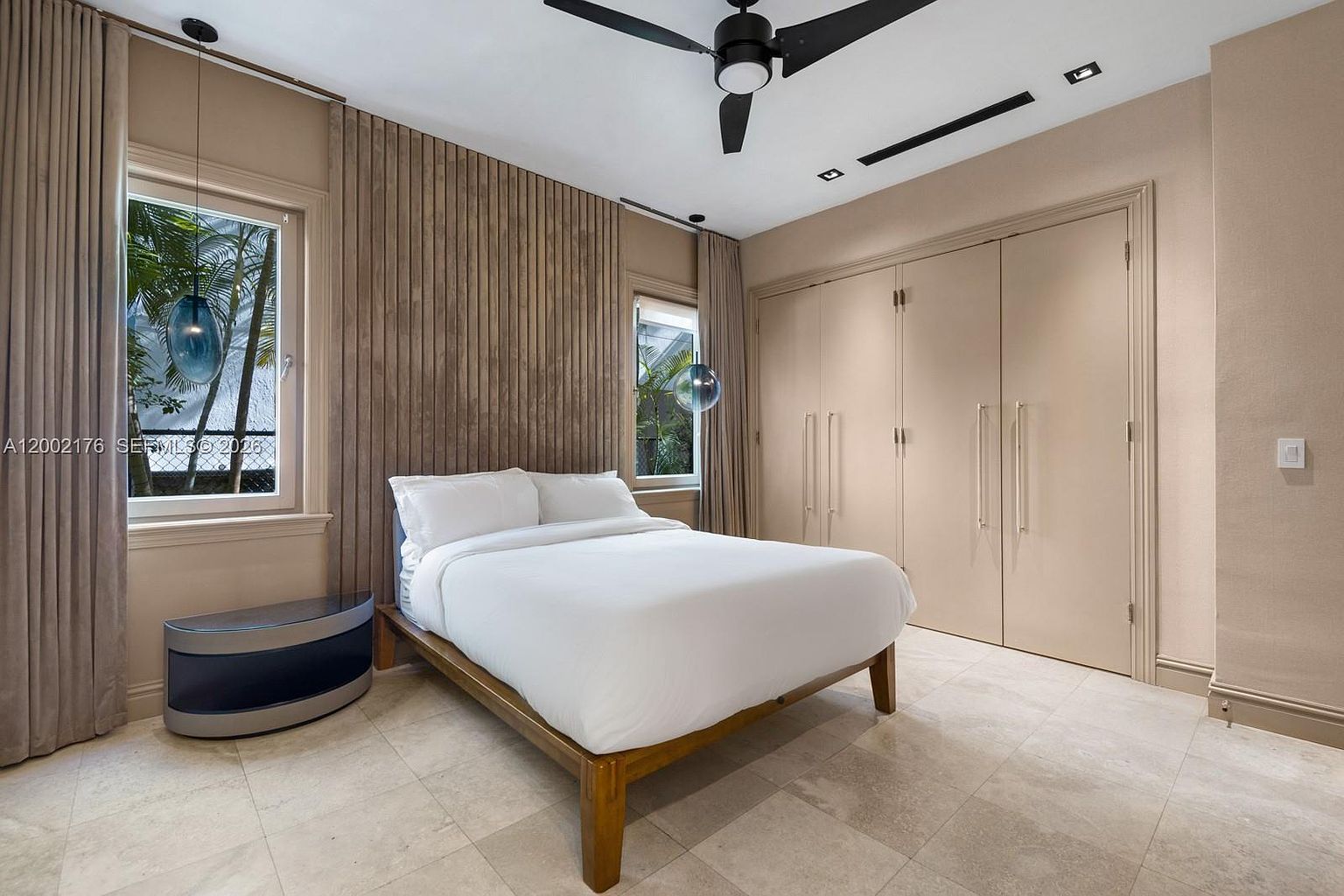 This contemporary bedroom features a platform bed centered against a textured, vertical-slat wood accent wall. The room is illuminated by two modern, spherical pendant lights hanging near the windows, which provide a view of lush greenery outside. A large, neutral-toned closet with tall doors occupies the right wall, while the space is finished with light-colored tile flooring and a ceiling fan.