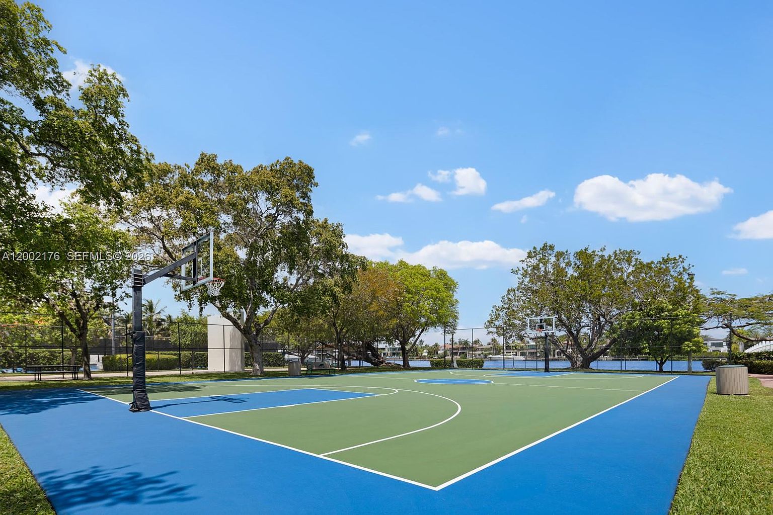 This image showcases a well-maintained outdoor basketball court situated in a scenic community setting. The court features a vibrant blue and green color scheme, framed by lush green trees and a calm body of water in the background. The perspective is from the court level, highlighting the recreational amenities available to residents.