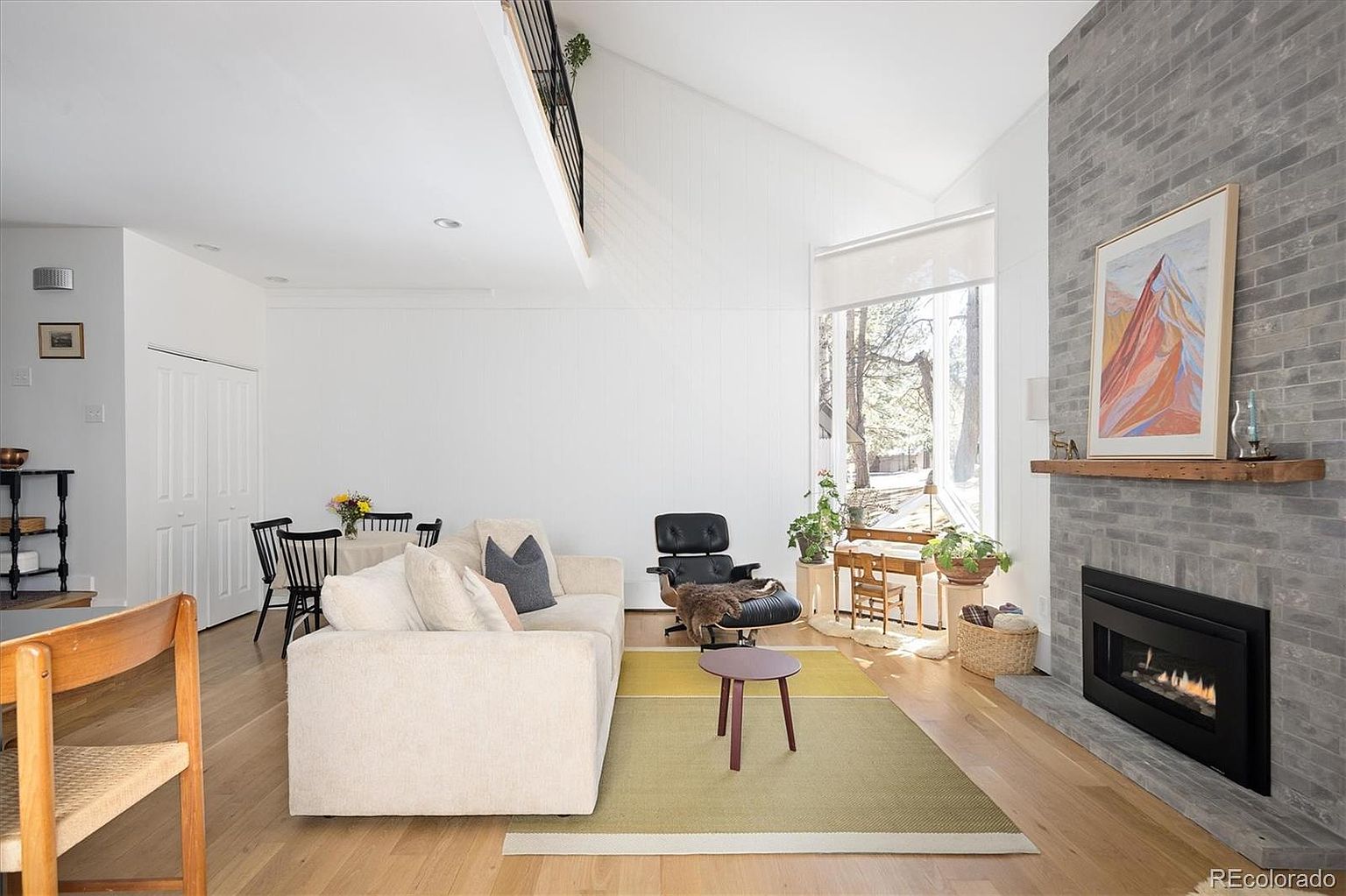 This bright and airy living room features a soaring vaulted ceiling and a striking floor-to-ceiling gray brick fireplace as its focal point. The space is furnished with a comfortable cream-colored sofa, a classic Eames-style lounge chair, and a minimalist area rug, all set against light hardwood floors. Large windows allow for abundant natural light, creating a warm and inviting atmosphere that seamlessly blends modern design with cozy, functional living.