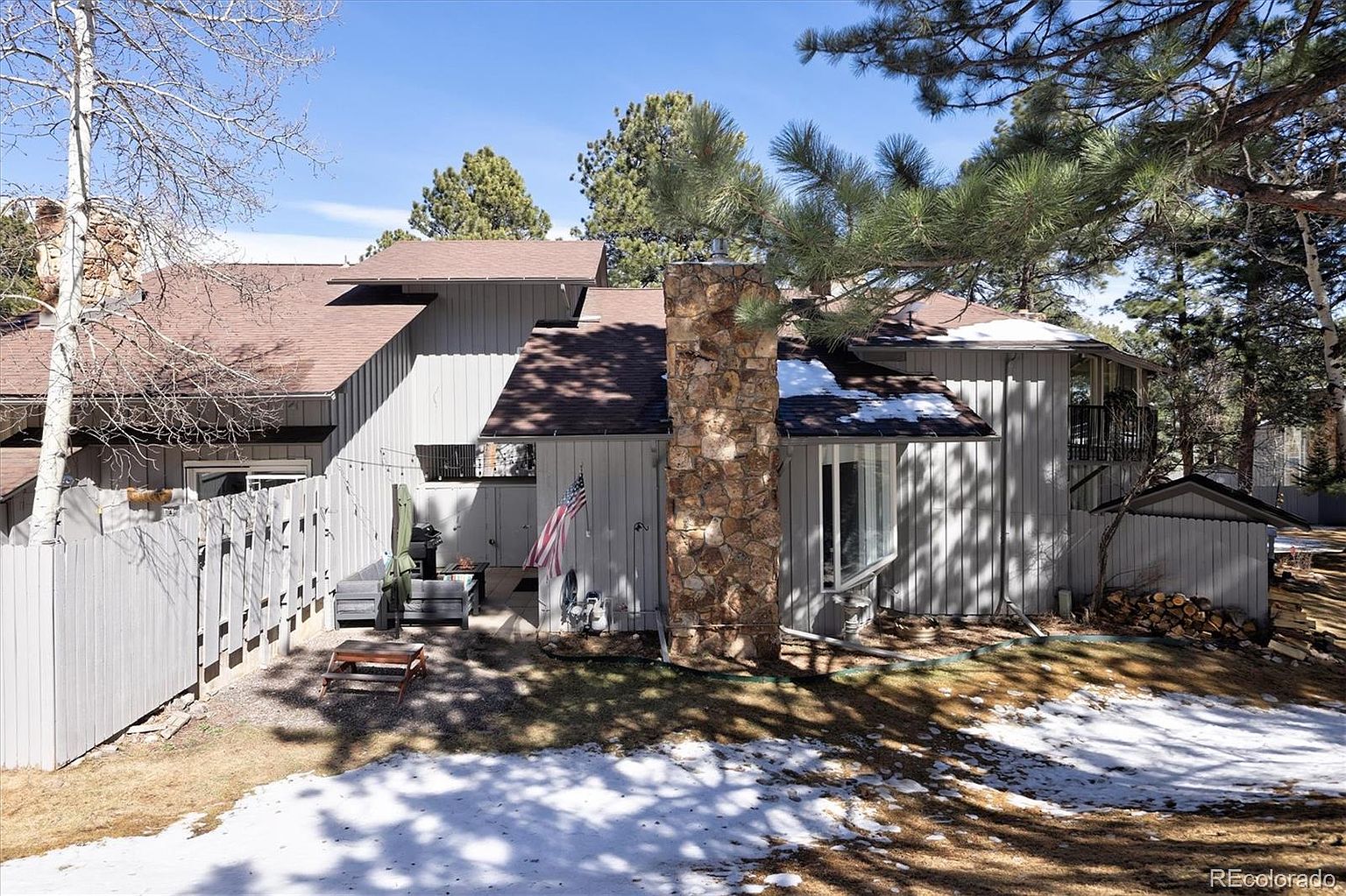 This rear view of a multi-level home showcases a rustic aesthetic with vertical wood siding and a prominent stone chimney. The property features a cozy patio area with outdoor seating, a small American flag, and a wood pile, all set against a backdrop of pine trees and patches of lingering snow. The perspective is captured from a slightly elevated angle, highlighting the home's integration into its wooded, mountainous environment.
