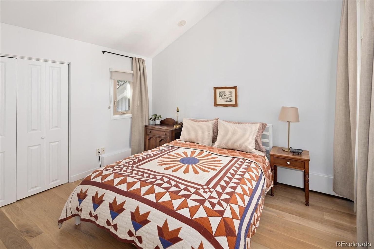 This cozy bedroom features a vaulted ceiling and light-toned hardwood flooring, creating an airy and inviting atmosphere. The room is centered around a bed with a vibrant, patterned quilt, flanked by two wooden nightstands and a simple table lamp. A window with neutral curtains allows natural light to brighten the space, while a white closet door provides clean, functional storage.