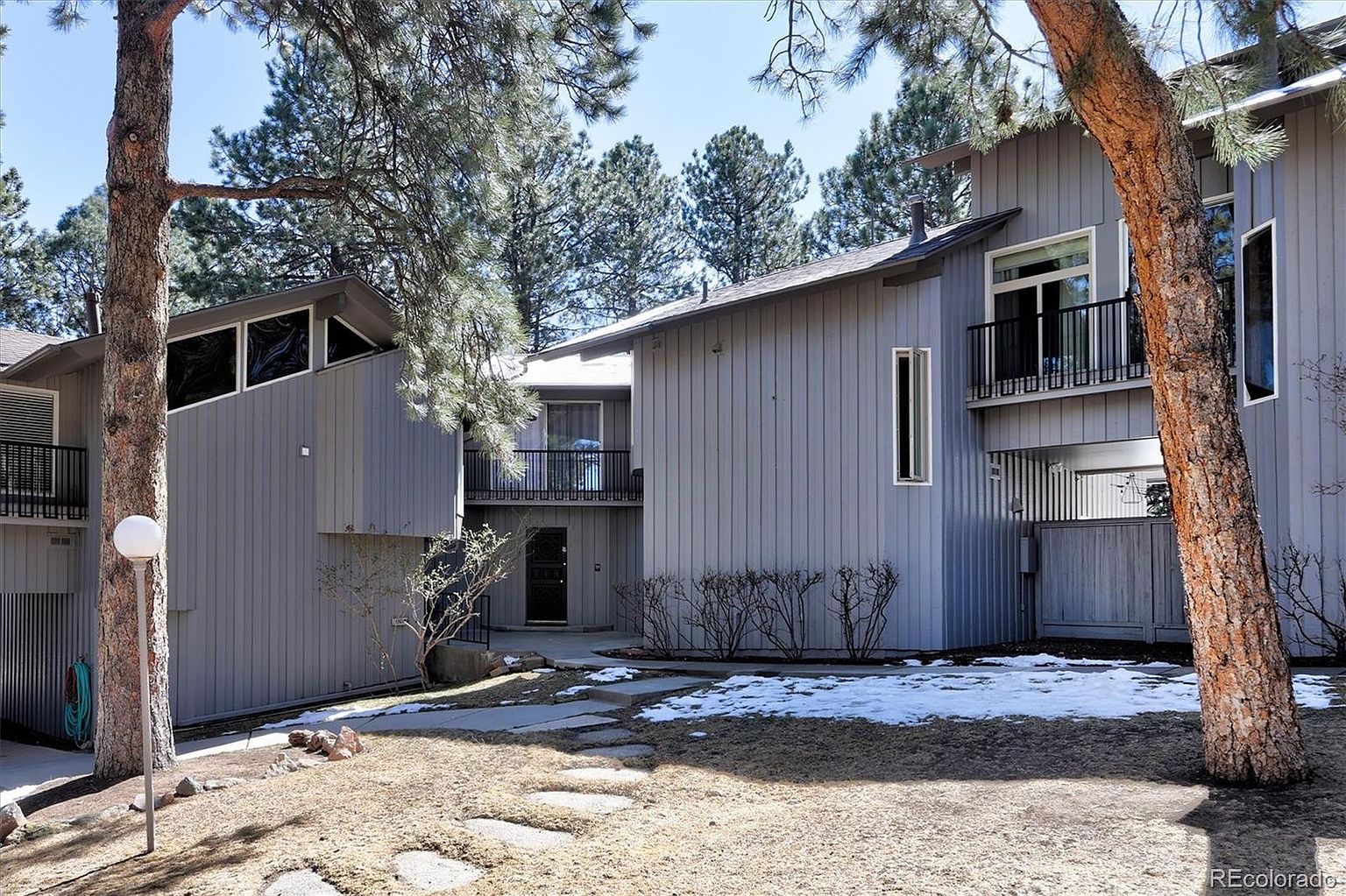 This exterior view showcases a multi-level, contemporary-style home with vertical gray siding and prominent pine trees framing the scene. A stone pathway leads through a partially snow-covered yard toward the recessed entryway, while balconies on the upper levels add architectural depth. The composition captures the home's unique geometric design and its integration into a wooded, natural landscape.