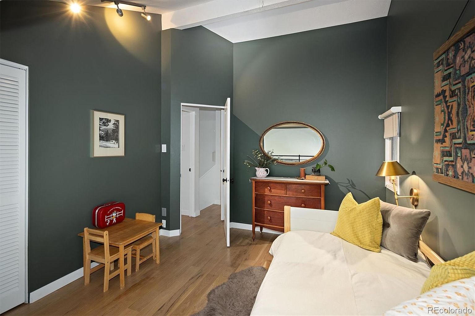 This cozy bedroom features dark, moody green walls contrasted by light wood flooring and a white daybed with vibrant yellow and grey accent pillows. A vintage wooden dresser topped with a mirror and decorative items sits against the wall, while a small child-sized table and chair set adds a playful touch to the corner. The room is illuminated by a warm wall-mounted lamp and overhead track lighting, creating an inviting and eclectic atmosphere.