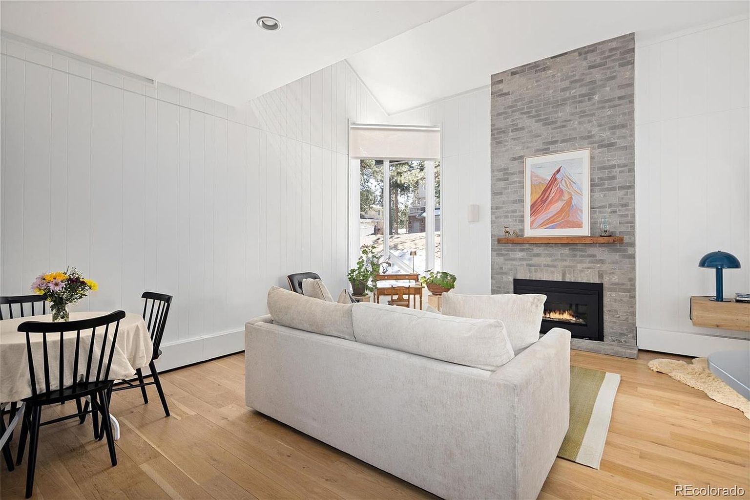 This bright and airy living room features a vaulted ceiling, light hardwood flooring, and a striking floor-to-ceiling gray brick fireplace as the central focal point. A comfortable, neutral-toned sofa faces the fireplace, while a small dining area with black spindle chairs is positioned to the left, creating an open-concept feel. Large windows allow natural light to flood the space, offering a pleasant view of the outdoors and contributing to a cozy, modern aesthetic.