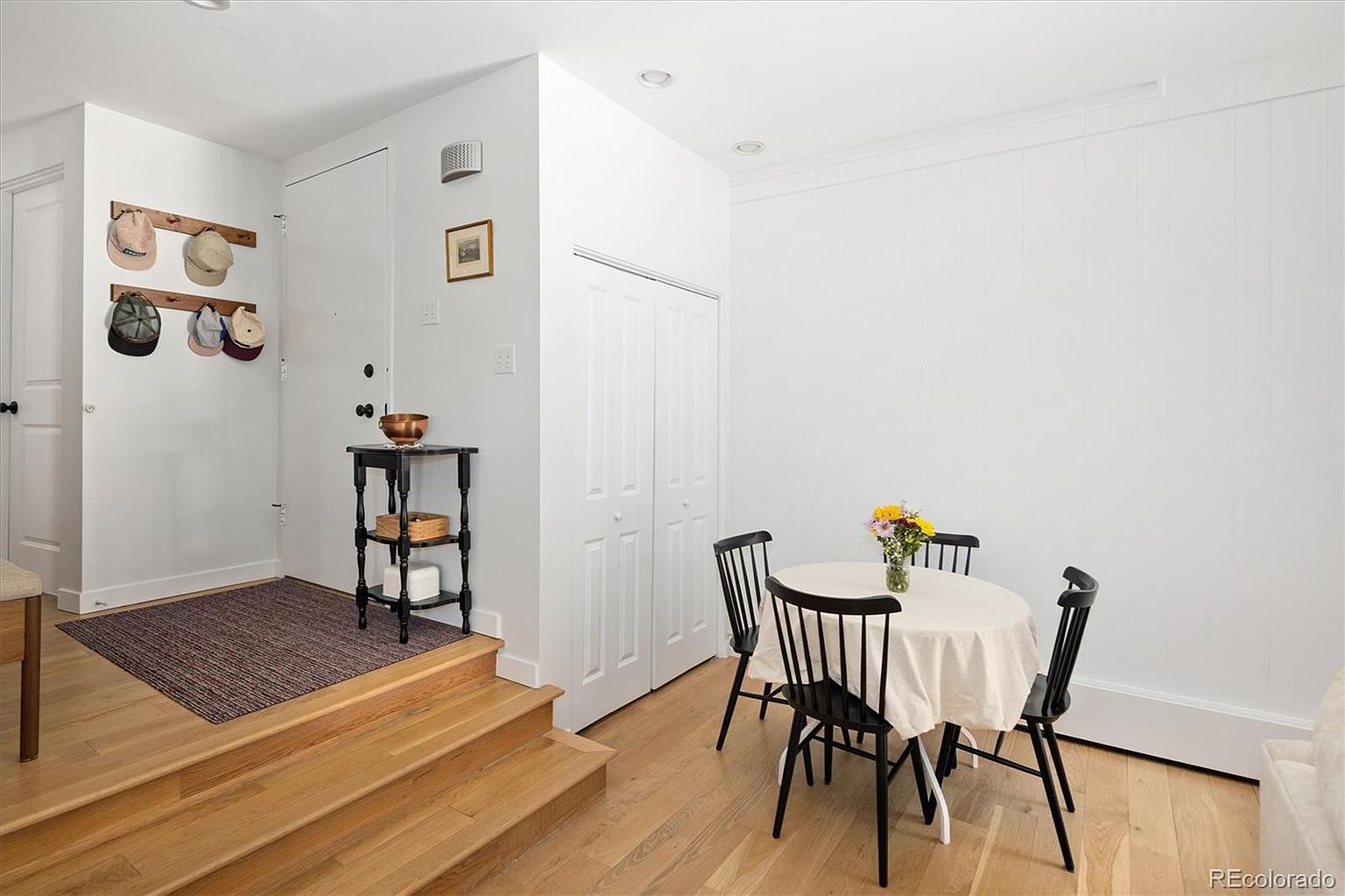 This bright and inviting dining area features a small, round table with a neutral tablecloth, surrounded by three black spindle-back chairs. The space is defined by light-toned hardwood flooring and a raised entryway platform with a dark wood console table and a hat rack. The clean white walls and minimalist aesthetic create a welcoming, airy atmosphere suitable for a cozy breakfast nook or casual dining space.