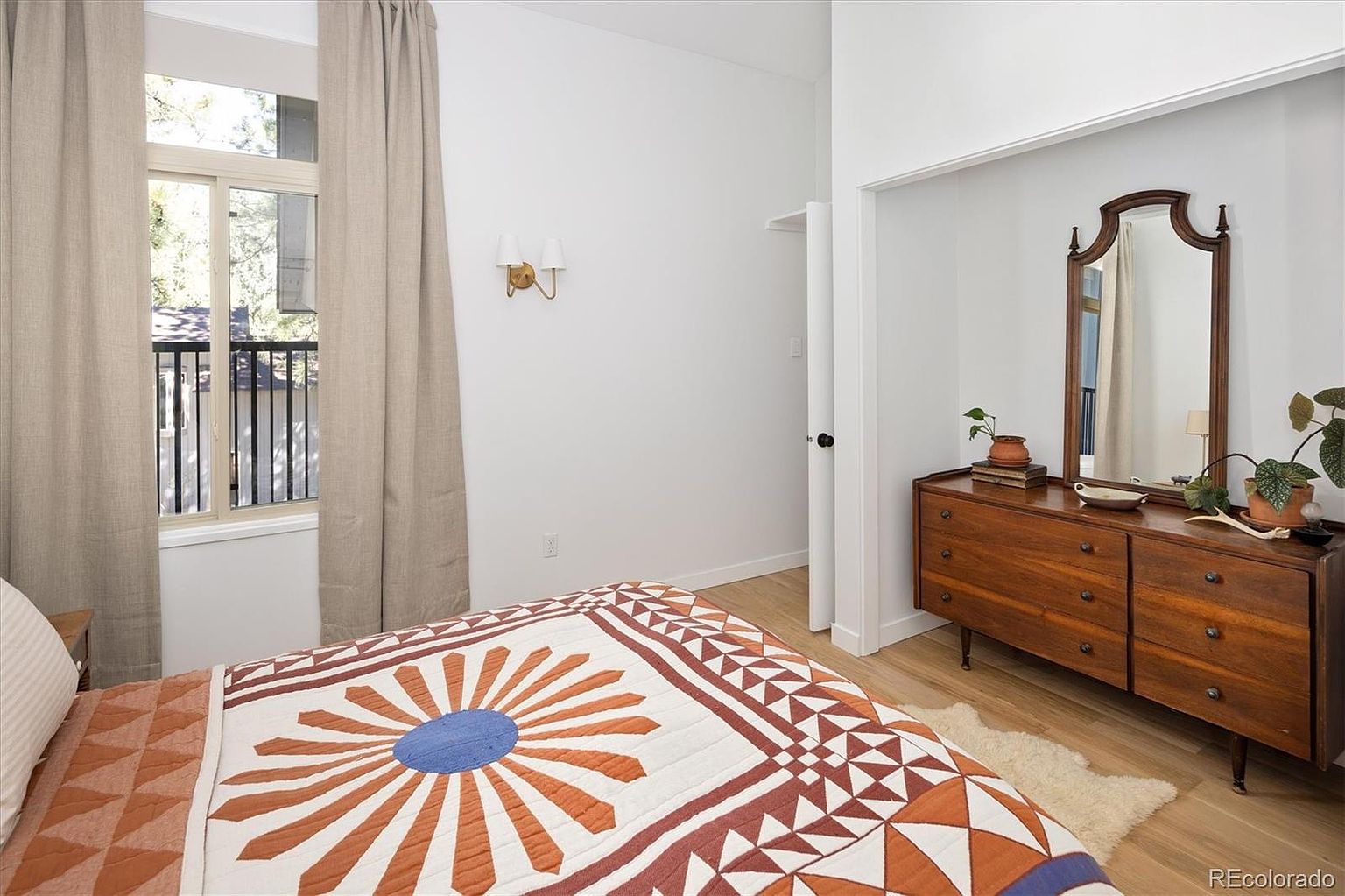 This cozy bedroom features a mid-century modern aesthetic, highlighted by a warm-toned wooden dresser topped with a vintage-style mirror and decorative plants. A patterned quilt with geometric orange and blue accents covers the bed, while soft, neutral curtains frame a window that looks out onto a balcony. The room is bright and airy, with light wood flooring and minimalist wall sconces, creating a welcoming and tranquil atmosphere.