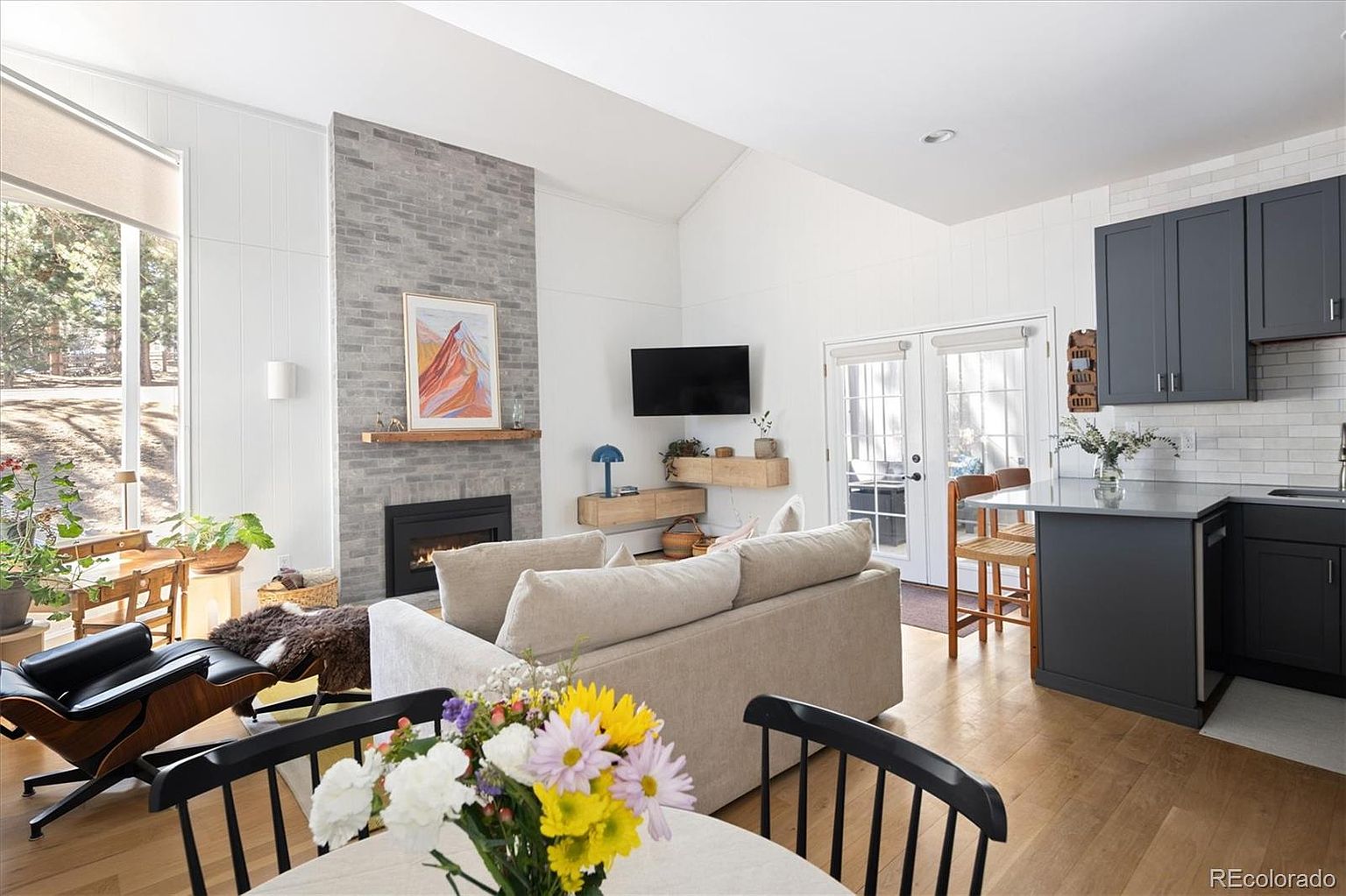 This open-concept living space features a striking floor-to-ceiling gray brick fireplace as a focal point, complemented by a cozy beige sofa and a modern lounge chair. The room flows seamlessly into a kitchen area with dark cabinetry and a breakfast bar, creating a bright and inviting atmosphere with high vaulted ceilings. The perspective is captured from the dining area, showcasing the cohesive design and natural light streaming through the large windows and glass doors.