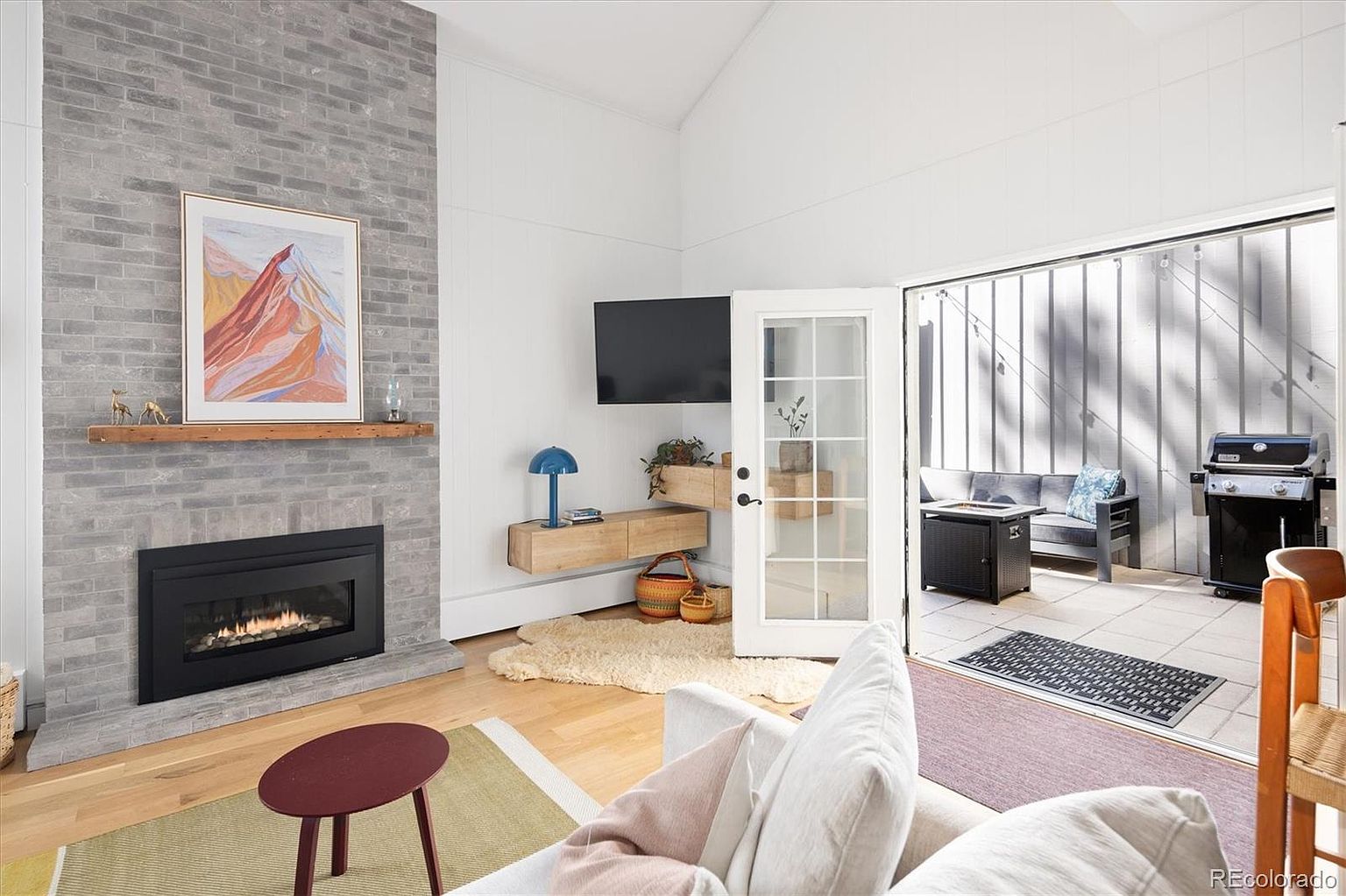This bright and airy living room features a striking floor-to-ceiling gray brick fireplace as its focal point, complemented by a natural wood mantel and a modern art piece. The space flows seamlessly to an outdoor patio through a set of French doors, creating an inviting indoor-outdoor living experience. The room is furnished with a comfortable sofa, a floating media console, and light hardwood flooring, offering a contemporary and cozy atmosphere.