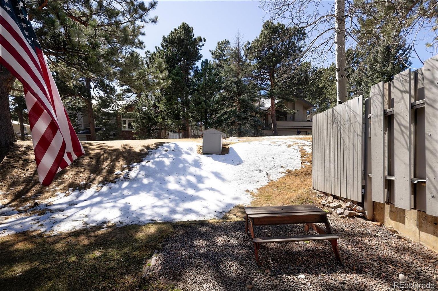 This backyard scene features a gravel patio area with a small wooden picnic table, bordered by a tall, light-colored wooden privacy fence. A gentle, snow-dusted grassy slope rises toward a backdrop of mature pine trees and a neighboring house, with an American flag visible in the foreground. The perspective is from a low angle, capturing the transition between the private outdoor living space and the natural, wooded surroundings.