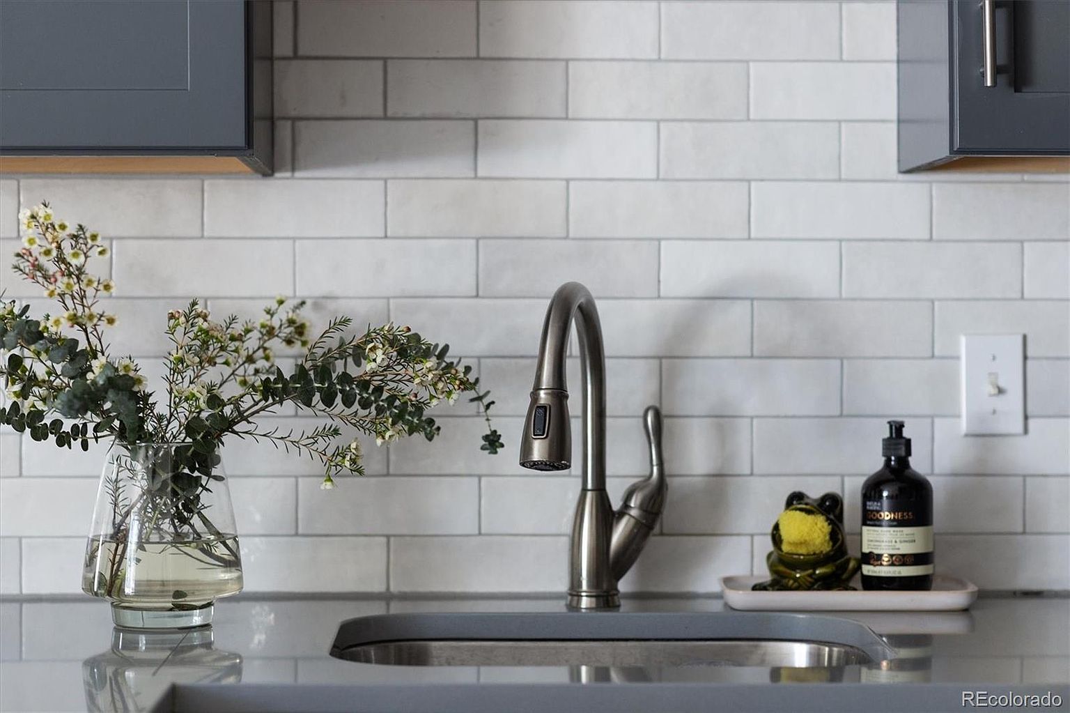 A close-up, eye-level shot of a modern kitchen sink area featuring a sleek, brushed-nickel pull-down faucet centered against a classic white subway tile backsplash. To the left, a clear glass vase holds a delicate arrangement of eucalyptus and small white flowers, while to the right, a whimsical frog-shaped sponge holder sits on a small tray next to a bottle of soap. The scene is clean and well-lit, highlighting the contemporary aesthetic and thoughtful decorative touches of the kitchen space.