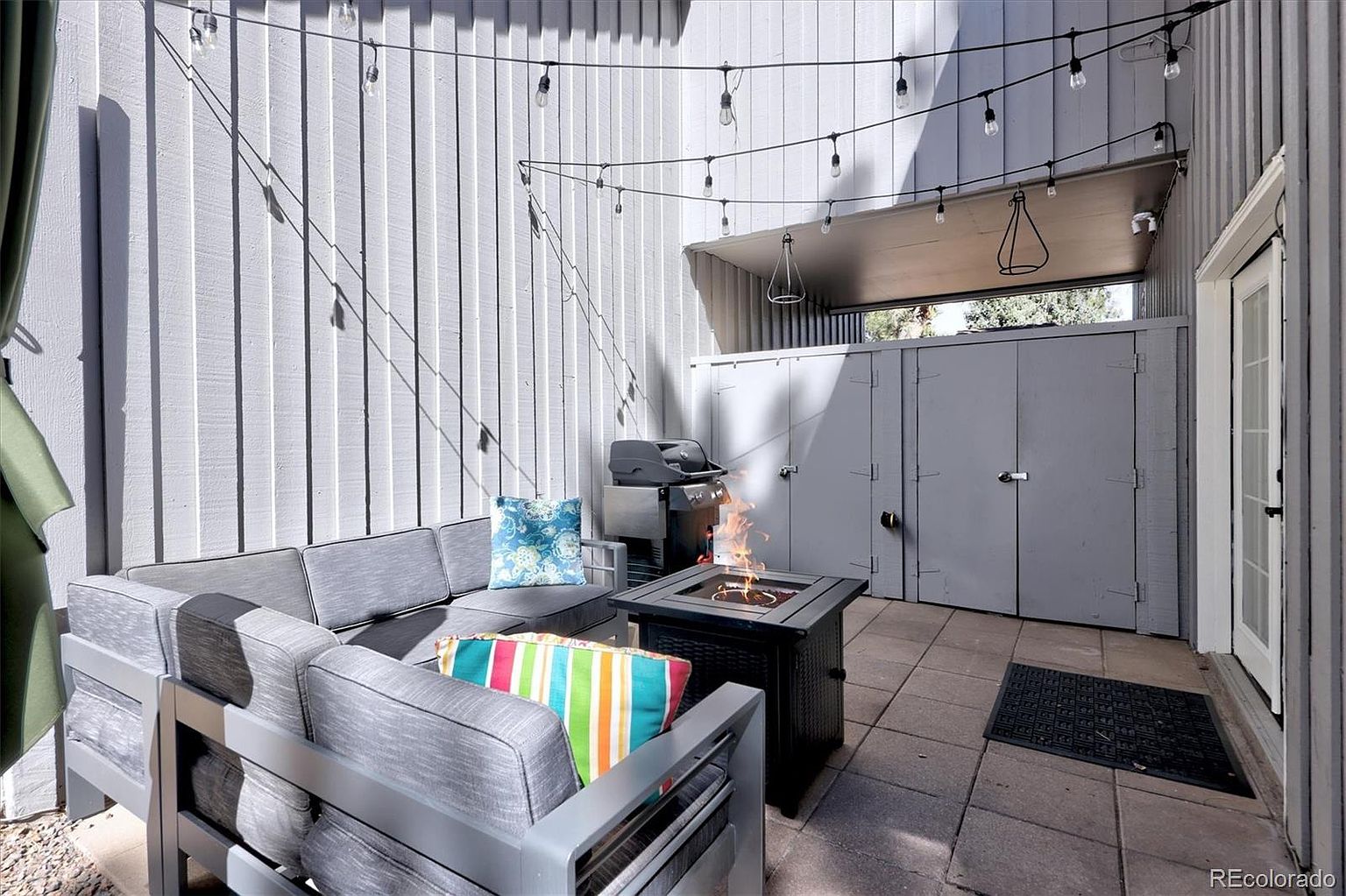 This cozy, private patio space features a modern grey sectional sofa adorned with colorful accent pillows, centered around a sleek black fire pit. The area is framed by vertical grey siding and illuminated by charming string lights, creating an inviting atmosphere for evening relaxation. A grill is tucked into the corner, and a door provides direct access to the home, making this an ideal outdoor living and entertaining area.