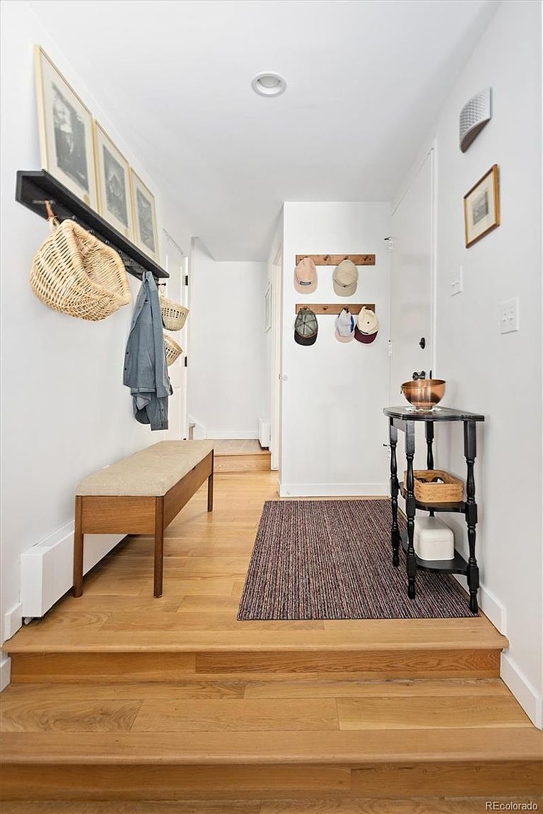 This bright and welcoming entryway features light hardwood flooring, a built-in wooden bench, and a black console table. The space is decorated with a wall-mounted coat rack, a shelf with framed art and a woven basket, and a textured area rug, creating a functional and stylish transition area. The perspective is a straight-on shot from the lower level looking up into the main hallway, emphasizing the clean, minimalist aesthetic.