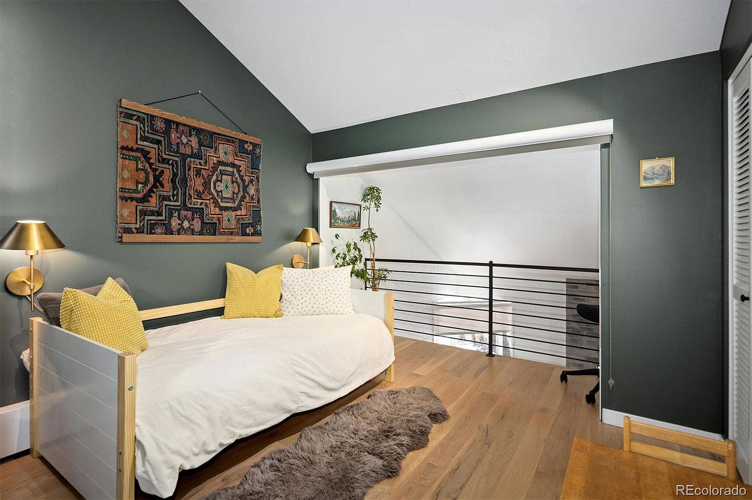 This cozy loft bedroom features a modern daybed with yellow accent pillows set against a striking dark green accent wall. The space is highlighted by a vaulted ceiling, light wood flooring, and a sleek black metal railing that overlooks the lower level. A decorative wall tapestry and warm wall-mounted lighting create an inviting and stylish atmosphere.