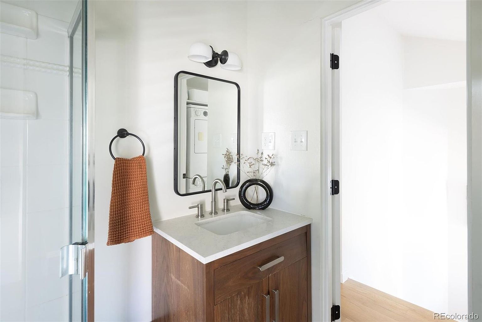 This modern bathroom vanity features a rich wood-toned cabinet topped with a sleek, light-colored stone countertop and an undermount sink. A rectangular black-framed mirror hangs above, complemented by a minimalist light fixture and a textured, rust-colored hand towel hanging on a circular wall ring. The clean white walls and contemporary hardware create a bright, sophisticated, and inviting atmosphere.