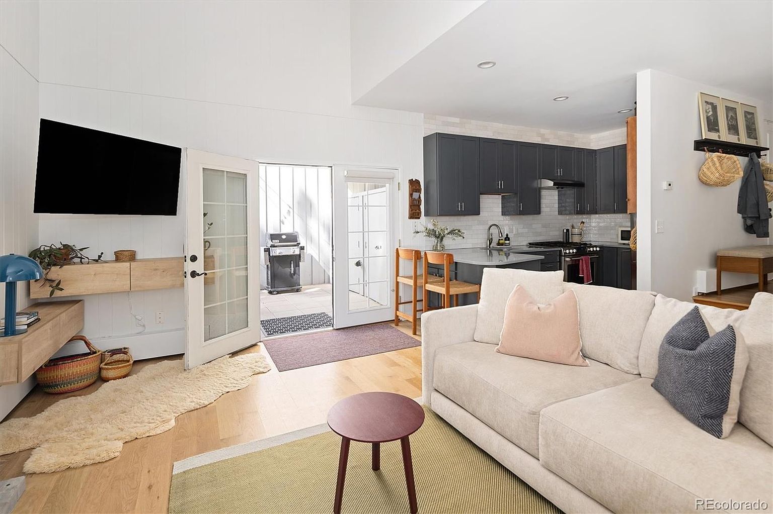 This bright and airy living room features a neutral-toned sofa, a modern circular coffee table, and light hardwood flooring, creating a welcoming and contemporary atmosphere. The space flows seamlessly into an open-concept kitchen with dark cabinetry and a breakfast bar, while double glass doors provide a direct view and access to a private outdoor patio. The high ceilings and clean lines enhance the sense of openness, making it an ideal space for both relaxation and entertaining.