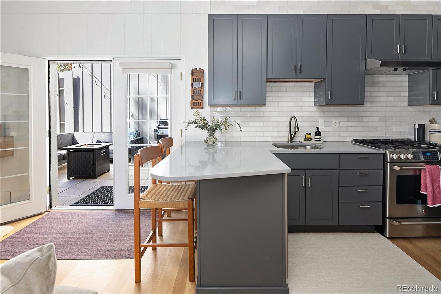 This modern kitchen features sleek dark gray cabinetry contrasted by a light gray quartz countertop and a classic white subway tile backsplash. The space includes a functional peninsula with bar seating, stainless steel appliances, and a seamless transition to an outdoor patio through glass doors. The overall aesthetic is contemporary and inviting, emphasizing a bright, open-concept layout.