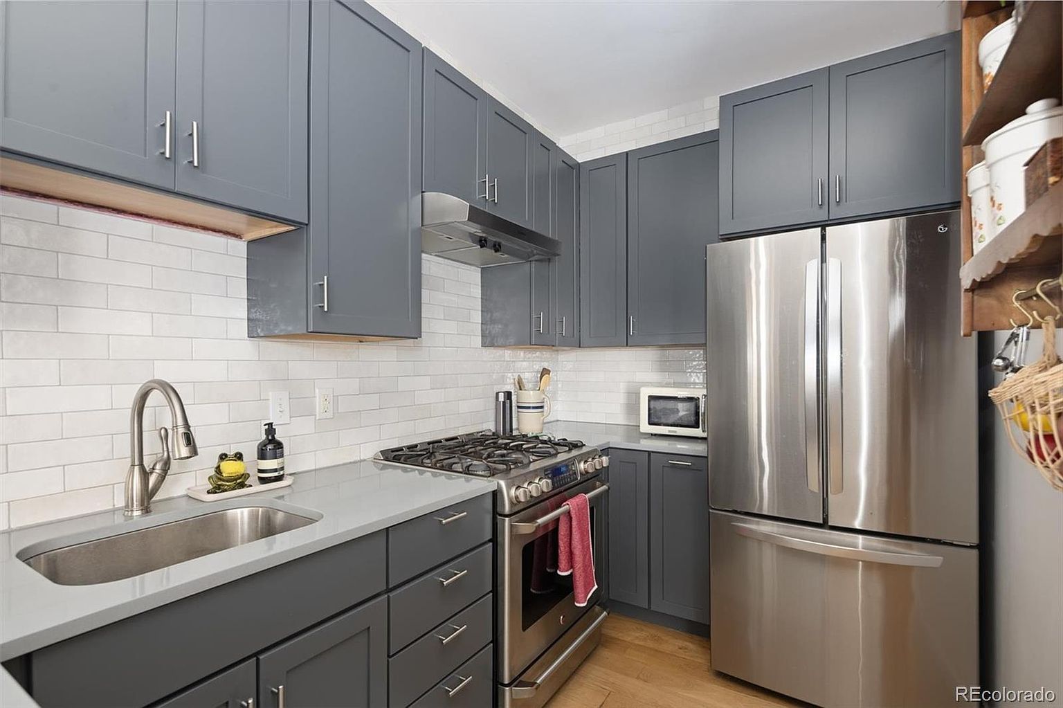 This modern, compact kitchen features sleek dark gray cabinetry paired with light gray quartz countertops and a classic white subway tile backsplash. The space is equipped with high-end stainless steel appliances, including a gas range and a full-size refrigerator, creating a functional and stylish culinary environment. The perspective is a straight-on eye-level shot that highlights the efficient layout and contemporary design aesthetic.