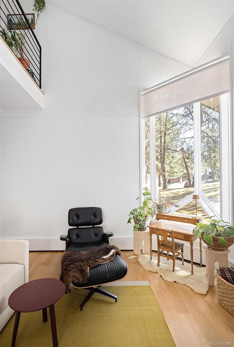This bright and airy living room features a high, vaulted ceiling and a large, multi-pane window that floods the space with natural light and offers a view of the outdoors. The room is furnished with a classic black leather lounge chair with an ottoman, a small wooden desk, and a modern side table, all set upon a textured yellow area rug. The clean white walls and light wood flooring create a minimalist, Scandinavian-inspired aesthetic that feels both cozy and sophisticated.